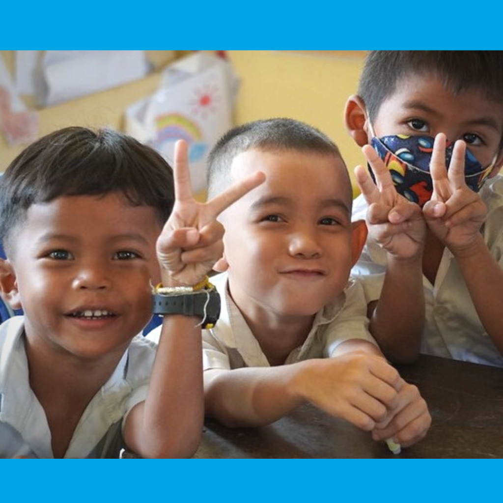 Three boys in school uniform smile and make peace signs at a school desk