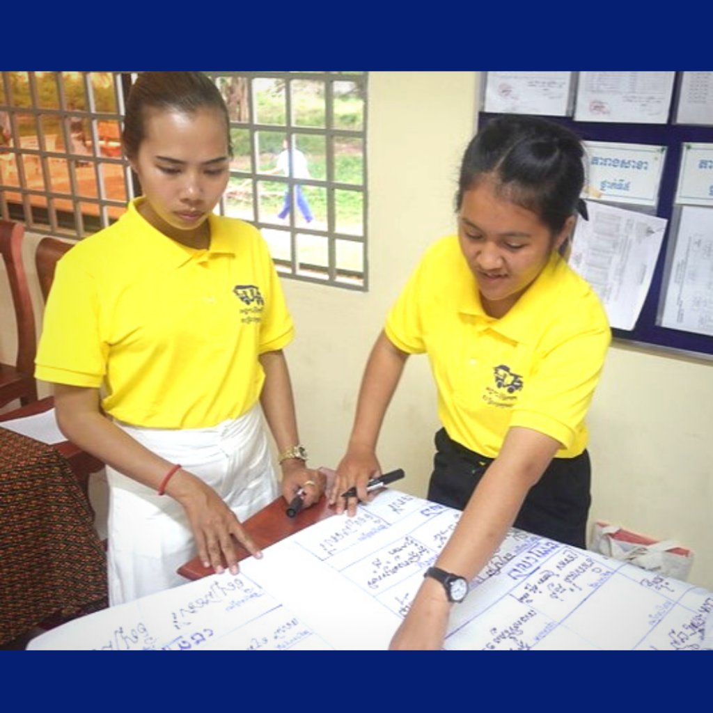 Two women in yellow TukTuk shirts write on large sheets of paper at a workshop table