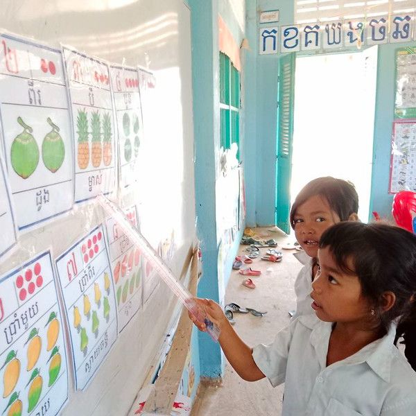 Two girls point at colourful classroom posters of fruit and numbers on a wall