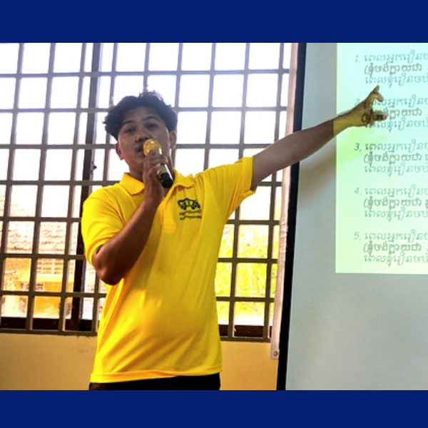 A TukTuk staff member in a yellow shirt speaks into a microphone and points at a projected slide