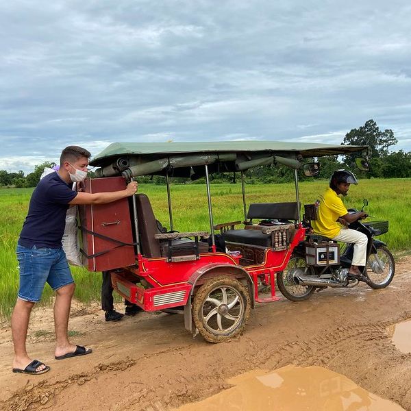 A man helps push a red tuk-tuk loaded with a cabinet along a muddy rural road