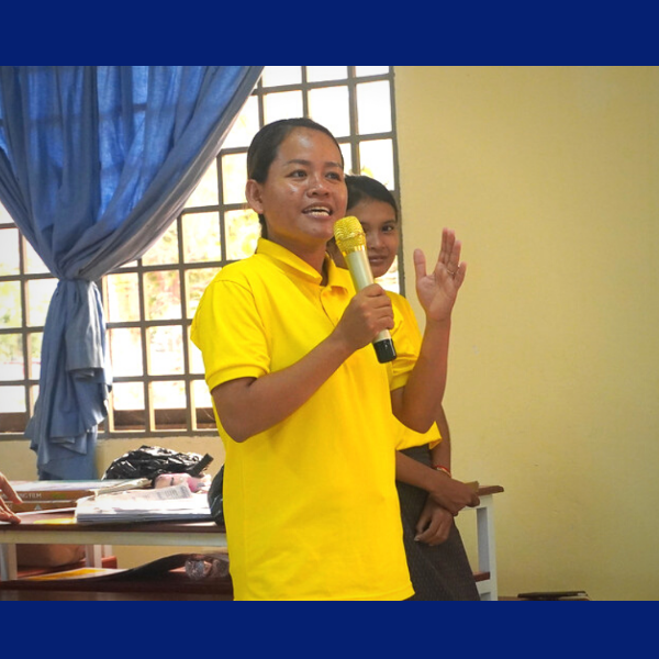 A teacher in a yellow shirt speaks into a microphone in front of a classroom curtain