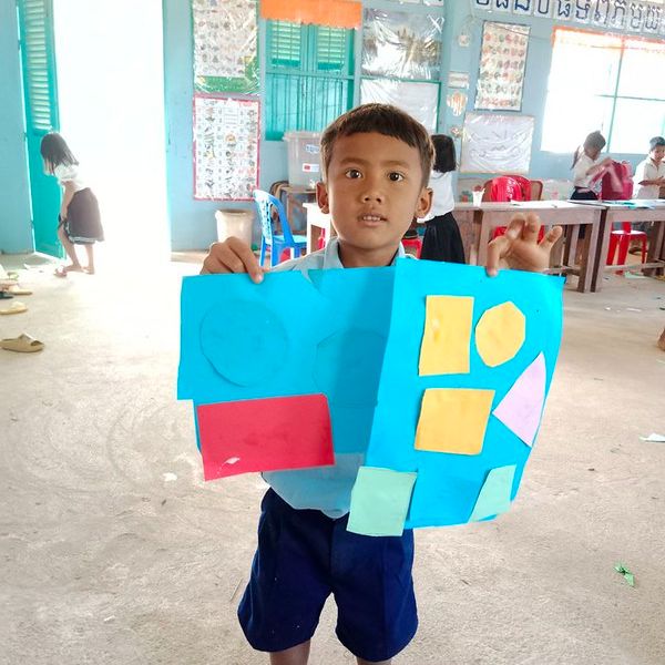 A boy holds up a blue paper collage of coloured shapes in a classroom