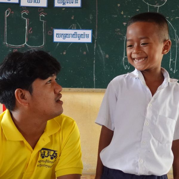 A TukTuk staff member in a yellow shirt sits beside a smiling boy at a chalkboard