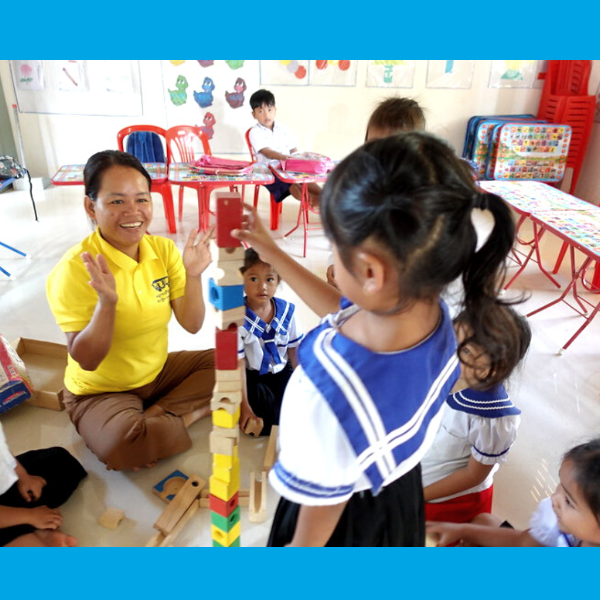 A woman in a yellow TukTuk shirt claps as a girl stacks wooden blocks into a tall tower