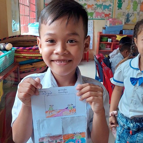 A smiling boy holds up a colourful drawing worksheet in a classroom