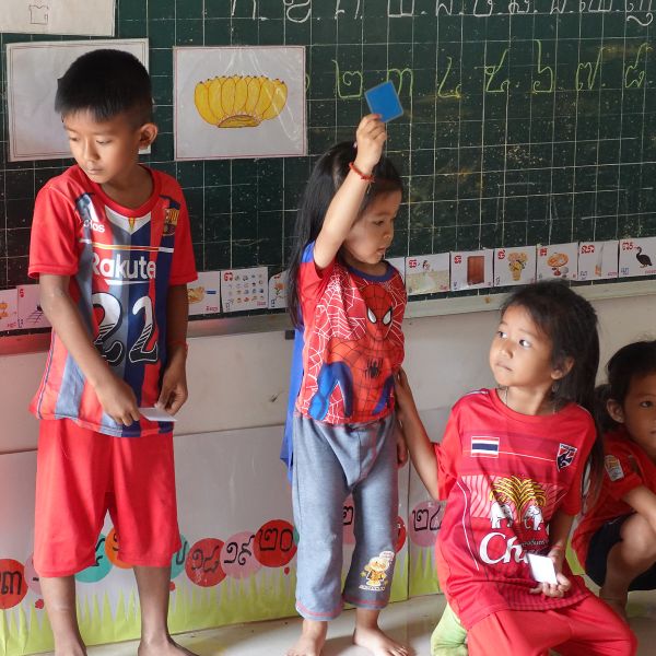 Children in red football shirts stand by a chalkboard, one holding up a blue card