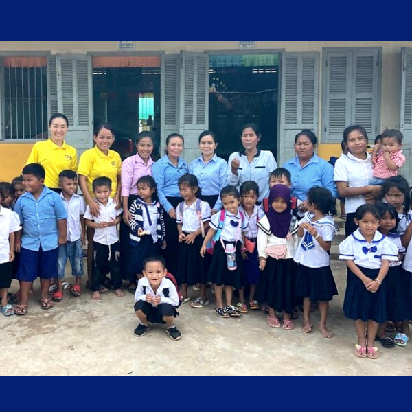 Teachers, TukTuk staff and many children in school uniform stand in front of a school building