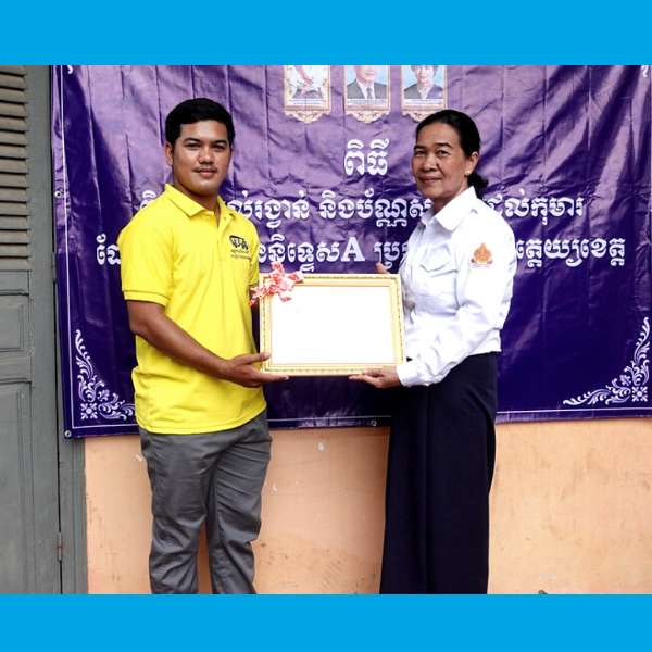 A TukTuk staff member and a woman in white hold a framed certificate in front of a banner