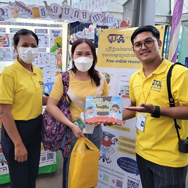 Two TukTuk staff in yellow shirts and a visitor show a Khmer picture book at the book fair booth