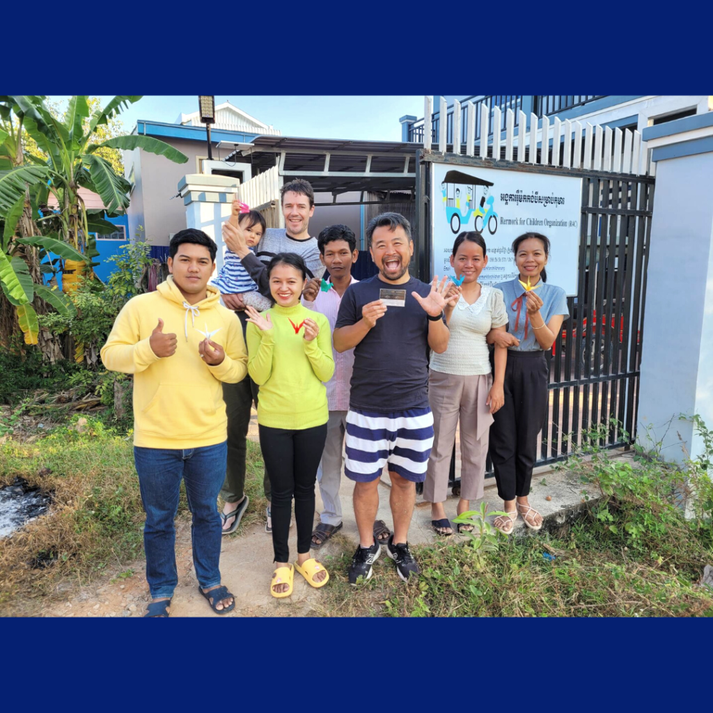 TukTuk staff and visiting volunteers pose outside the office gate, some giving a thumbs up