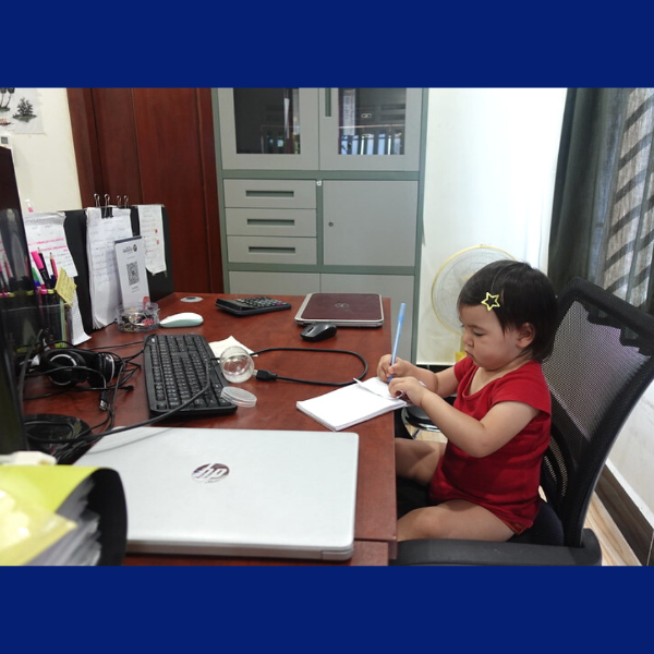 A toddler in a red top sits at an office desk with a notebook beside a laptop