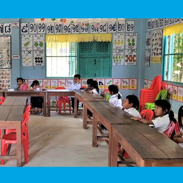 Children sit at long wooden tables in a classroom decorated with number and fruit posters