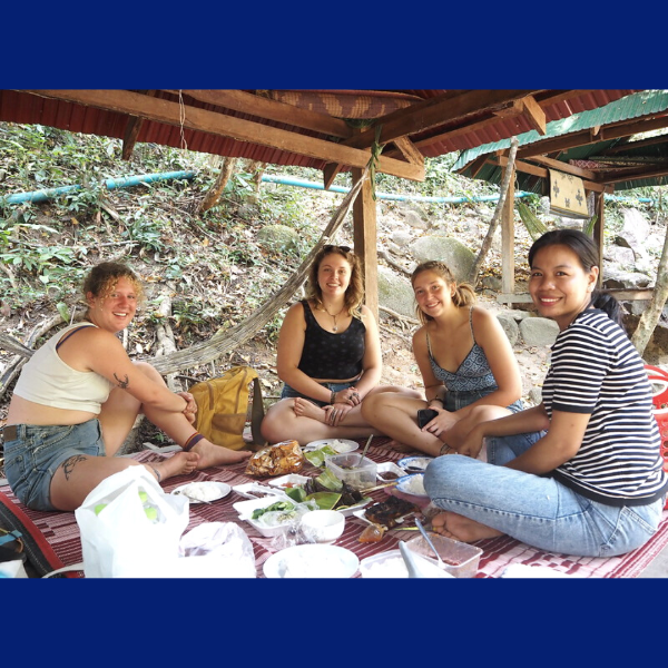 Voluntukers and a TukTuk staff member relax on a picnic mat under a wooden shelter
