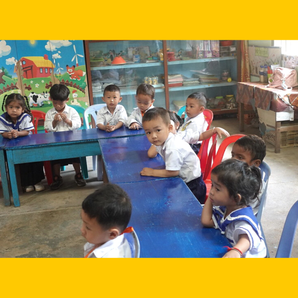 Young children in school uniforms sit at blue tables in a preschool classroom