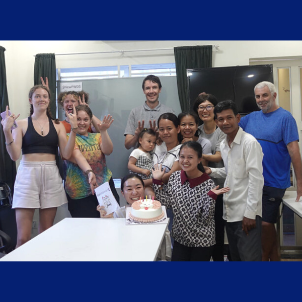 Voluntukers and TukTuk staff gather around a birthday cake in the office, waving at the camera
