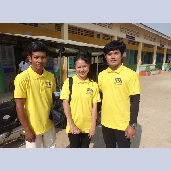 Three TukTuk staff members pose together in front of a school building during a follow-up visit