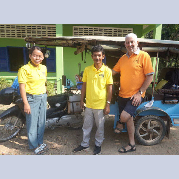 Two TukTuk staff and a visitor stand beside the light blue tuk-tuk in front of a school building