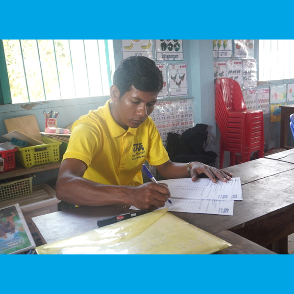 A TukTuk staff member fills in a form at a wooden desk in a classroom