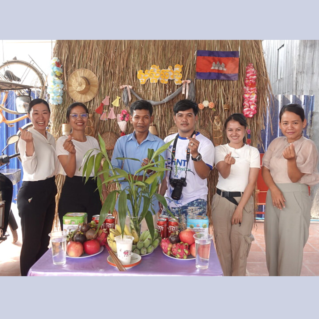 TukTuk staff and visitors pose in front of a straw wall with a Cambodian flag and a table of fruit