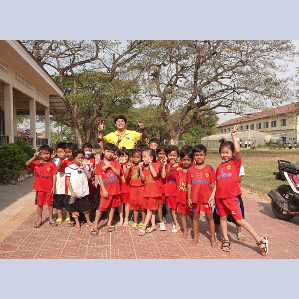 Children in red sports shirts pose outdoors at school, with a smiling TukTuk staff member behind them