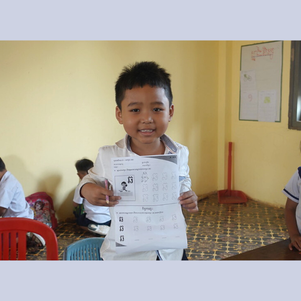 A schoolboy holds up a Khmer letter worksheet and smiles in his classroom