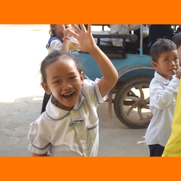 A smiling schoolgirl waves at the camera beside the light blue tuk-tuk