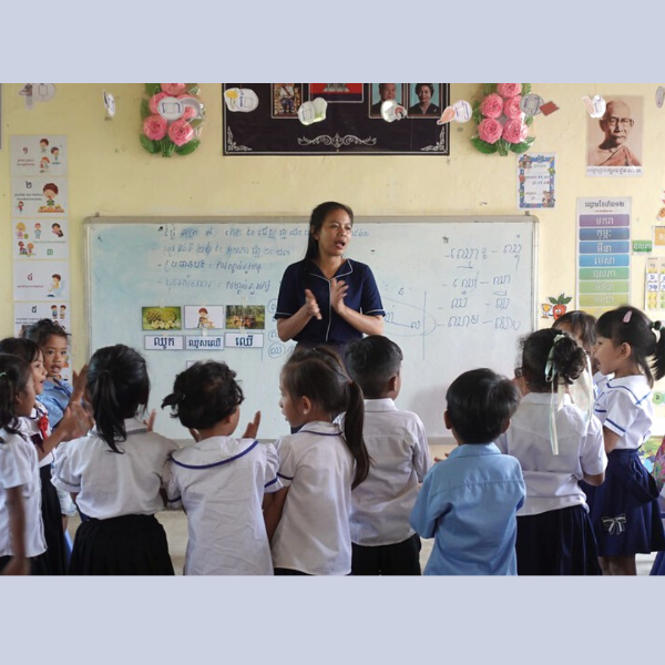 A teacher stands at a whiteboard in a decorated classroom while preschool children gather around to listen