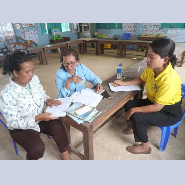 A TukTuk staff member takes notes while two teachers look through papers and books at a classroom table