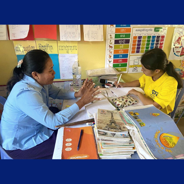 Two women sit at a desk piled with books and notebooks, talking while one takes notes