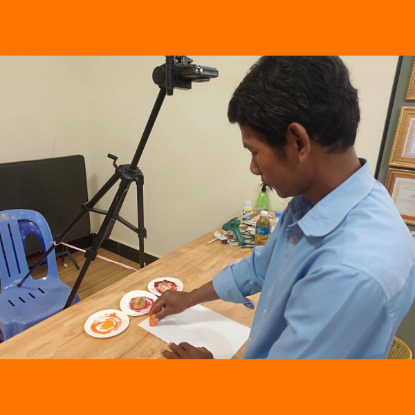 A TukTuk staff member prepares craft materials on painted paper plates at a table, with a camera on a tripod