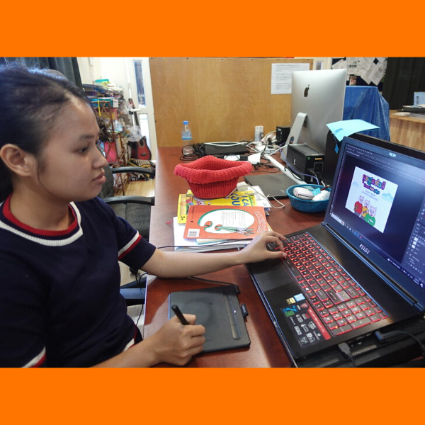 A young woman on the TukTuk team designs on a laptop with a drawing tablet at a crowded desk
