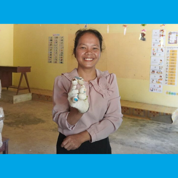 A smiling teacher holds a small hand puppet in an empty classroom