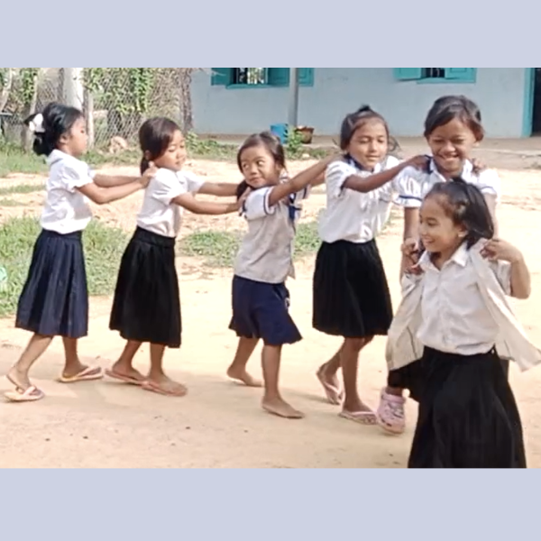 Schoolgirls in white blouses and black skirts hold each other's shoulders and dance in a line outdoors
