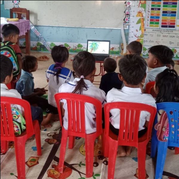 Preschool children sit on red chairs in a row watching a video on a laptop in class