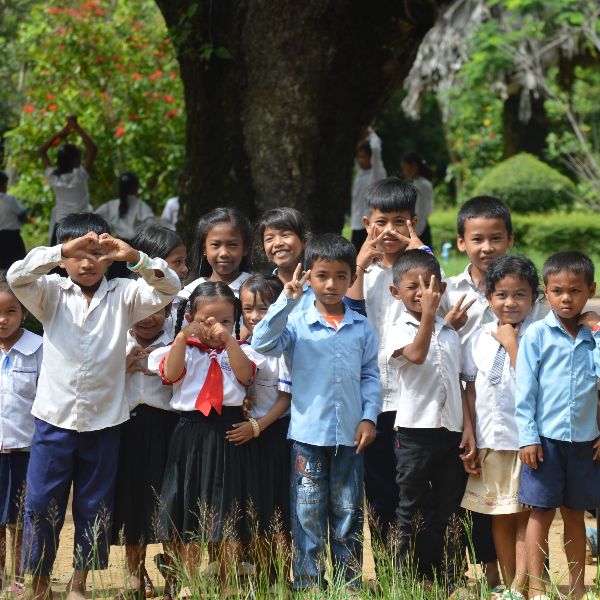 Schoolchildren in uniforms pose under a big tree, making heart shapes and peace signs with their hands