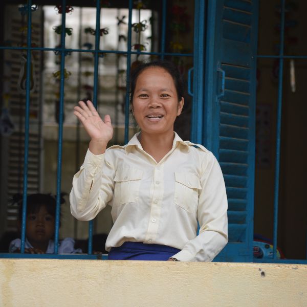 A teacher smiles and waves from a classroom window with blue shutters