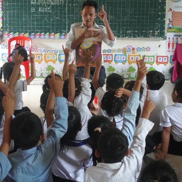 A teacher reads from a book at the board while schoolchildren raise their hands to answer