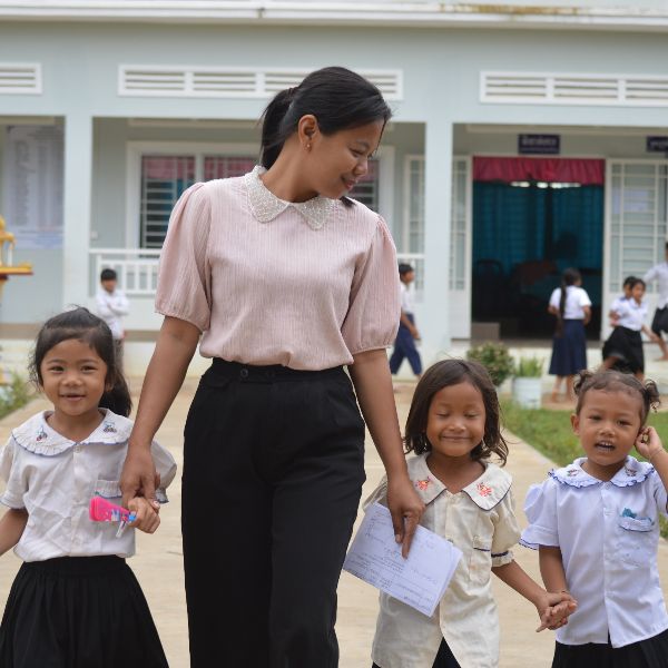 A woman walks hand in hand with three smiling preschool girls along a path in front of a school building