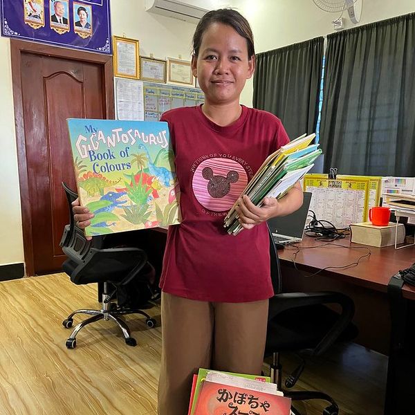 A TukTuk team member holds up a dinosaur Book of Colours and a pile of donated picture books
