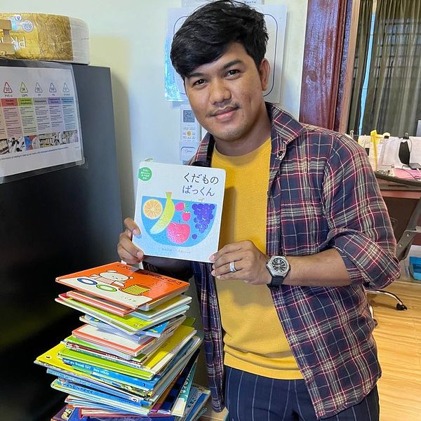 A man holds up a Japanese picture book beside a tall stack of donated picture books in the office