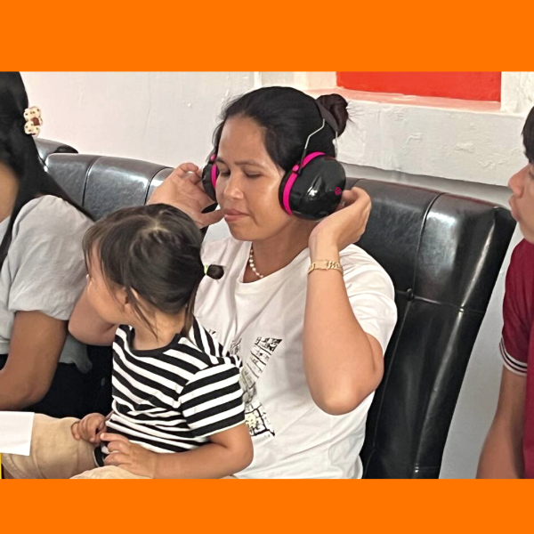 A woman wearing pink noise-reducing headphones holds a toddler on her lap on a sofa