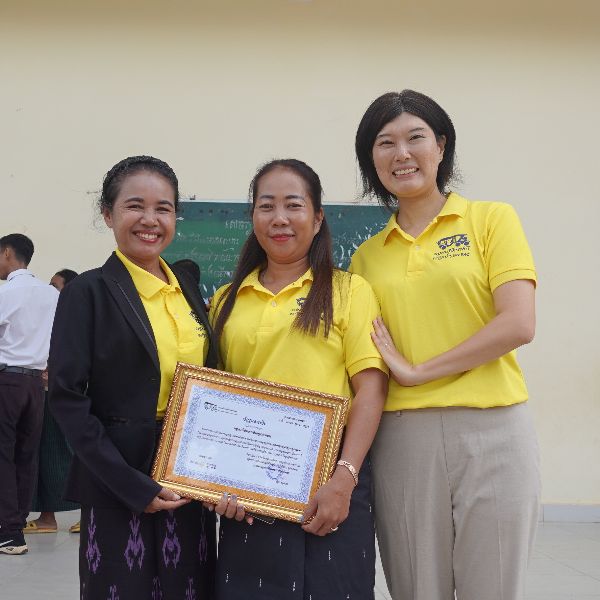 Two teachers with a framed certificate and a TukTuk staff member pose together in front of a green board