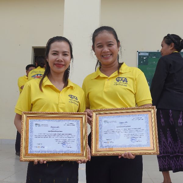 Two smiling teachers in yellow shirts hold framed certificates of completion