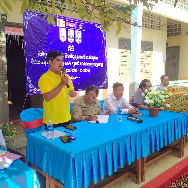 A TukTuk staff member speaks into a microphone beside officials seated at a table with a blue cloth