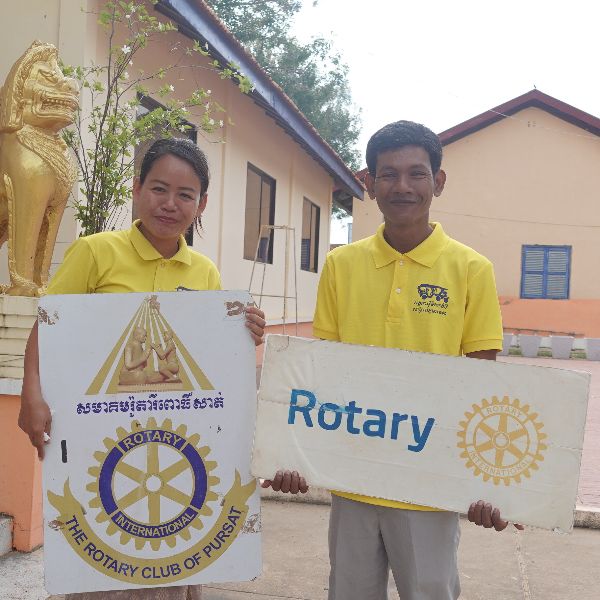 Two people in yellow shirts hold up Rotary Club of Pursat signs outside a school building