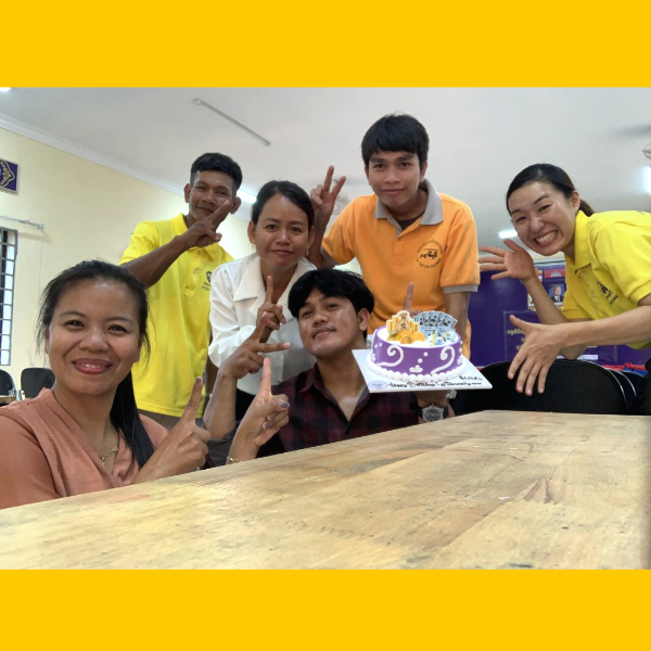 TukTuk staff and colleagues make peace signs around a purple birthday cake at a long table