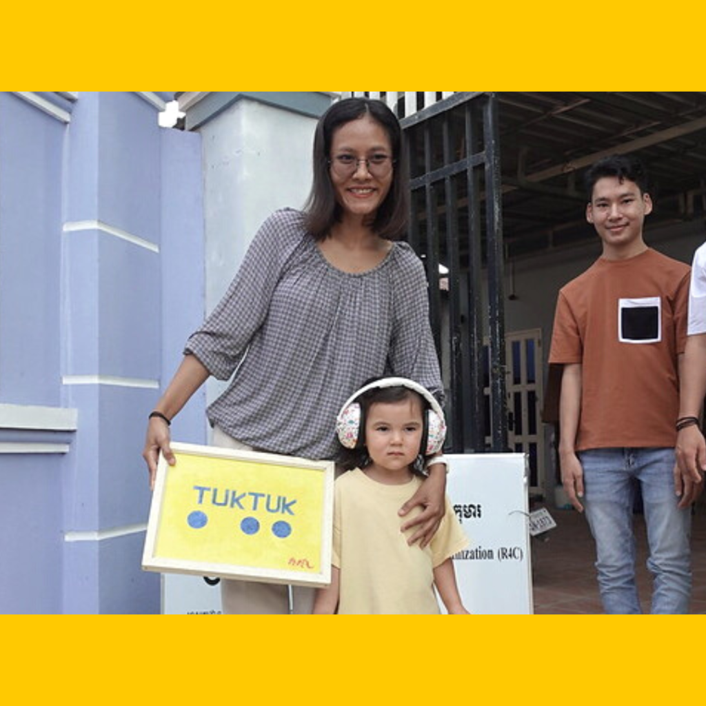 A woman and a young man stand with a toddler in pink headphones holding a TukTuk sign outside the office