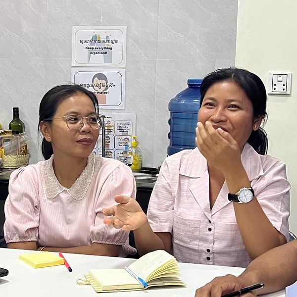 Two TukTuk staff members talk and laugh at a table with notebooks in the office