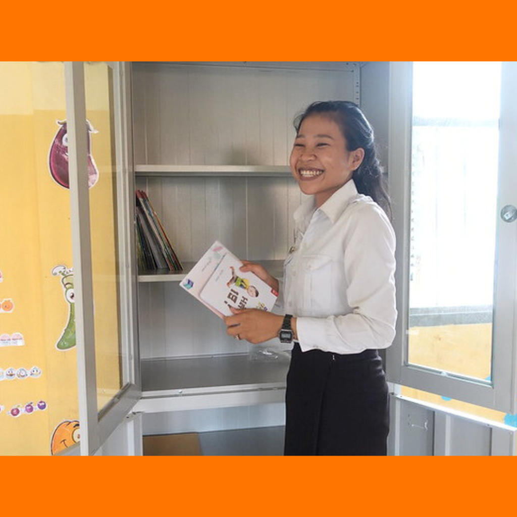 A teacher smiles and holds a book beside a newly installed metal cabinet in her classroom